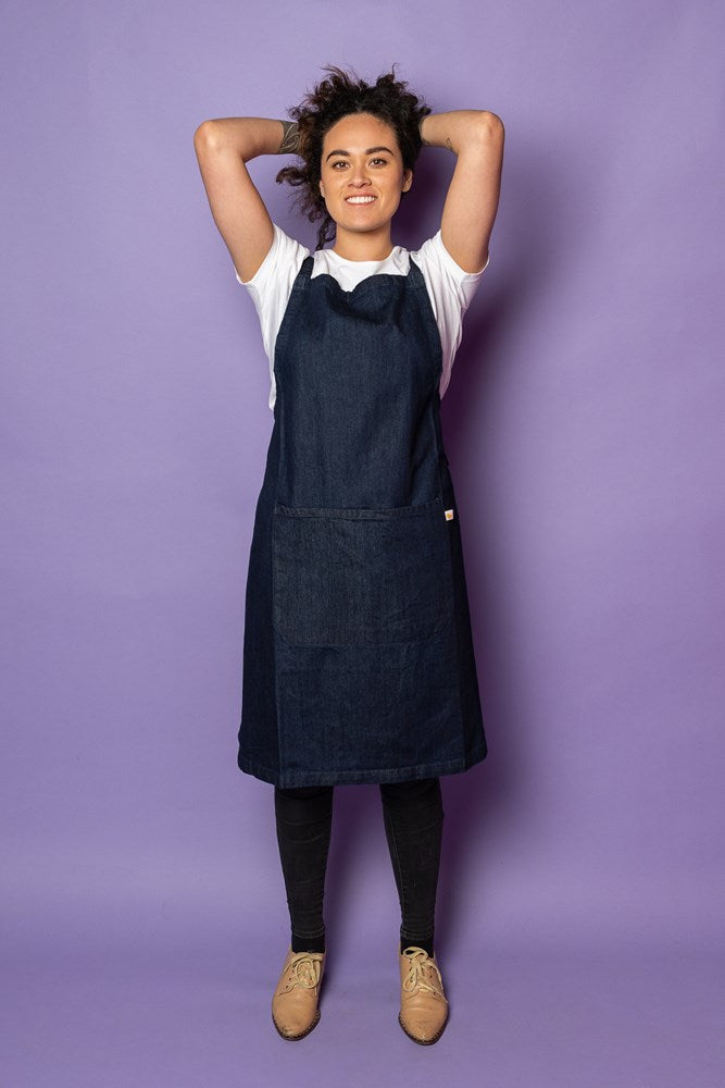 Female model in studio with hands behind head wearing Denim Fearless Apron against purple background